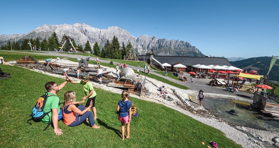 Familien genießen den Wasserspielplatz an der Karbachalm vor imposanter Hochkönig-Kulisse bei Sonnenschein