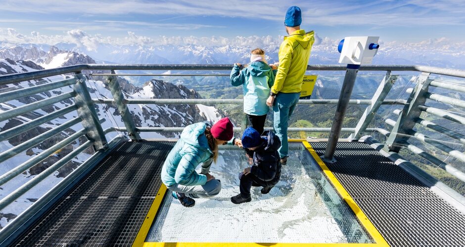 Familie steht auf dem Sky Walk mit Glasboden am Dachstein und blickt auf verschneite Berge und grüne Täler. | © Harald Steiner