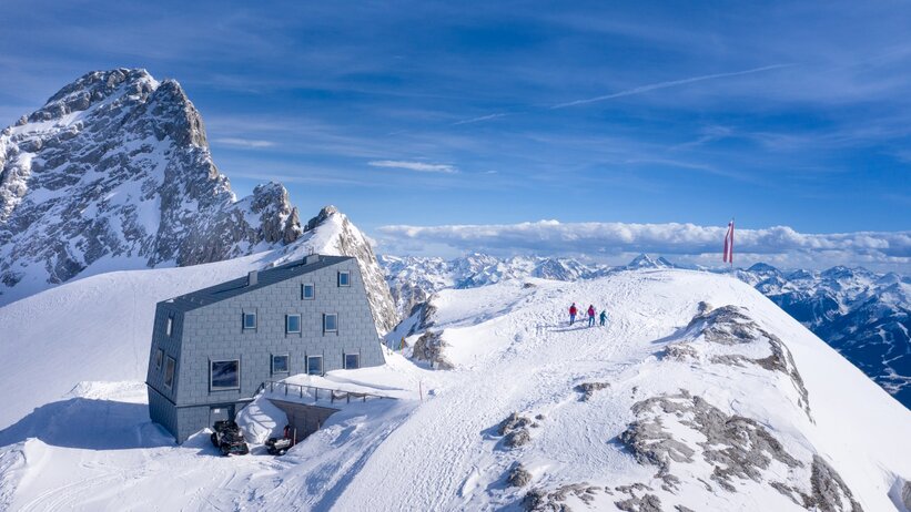 Moderne Berghütte auf schneebedecktem Gipfel mit Panorama, drei Personen wandern zur Spitze, österreichische Fahne im Wind. | © Josh Absenger
