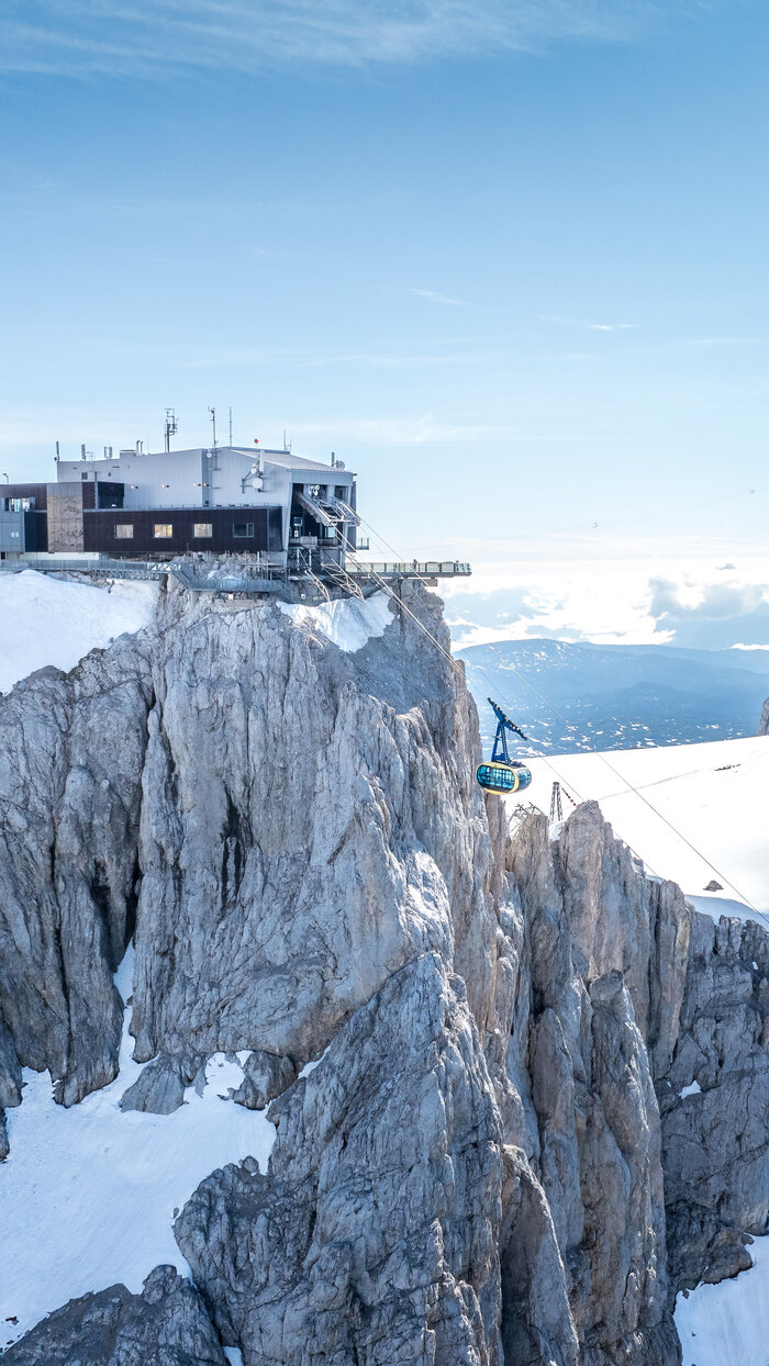 Cable car to mountain station on rugged cliffs | © Josh Absenger A blue cable car ascends to a modern mountain station on rugged cliffs, offering a wide view of snow-covered peaks and valleys. | © Josh Absenger