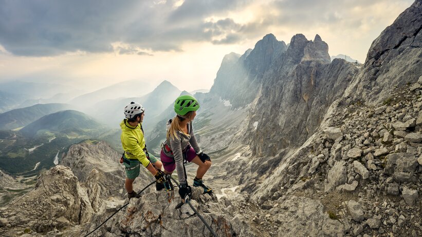 Zwei Bergsteiger mit Helmen und Kletterausrüstung auf Felsgrat mit Blick auf die markanten Gipfel des Dachsteinmassivs bei Sonnenuntergang. | © Peter Burgstaller