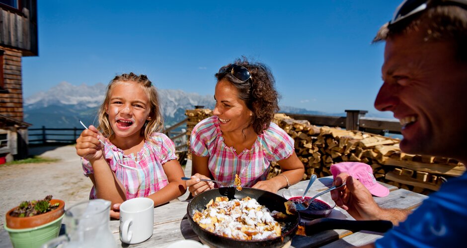 Familie isst Kaiserschmarrn auf Sonnenterrasse mit Almhüttenflair und Blick auf die Berge der Hochwurzen. | © Tom Lamm