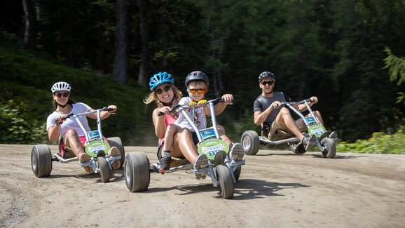 Drei Erwachsene und ein Kind fahren lachend mit Mountain-GoKarts auf Waldweg bei sonnigem Wetter. | © Harald Steiner