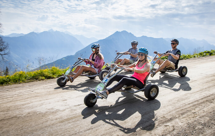 Mountain go-kart descent on Hochwurzen in sunshine | © Harald Steiner Four people ride mountain go-karts downhill on gravel road with alpine views and a summer vibe. | © Harald Steiner