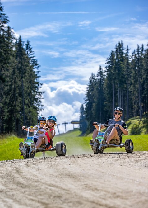 Zwei Erwachsene und ein Kind fahren mit Mountain-GoKarts über einen Schotterweg zwischen Wald und Wiese. | © Harald Steiner
