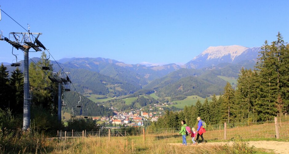 Familie wandert entlang eines Wiesenwegs mit Blick auf Berge, Wälder, Sessellift und das Tal mit Dorf. | © Katharina Wassler