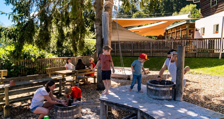 Kinder spielen barfuß mit Wasser und Holzfässern im schattigen Spielplatzbereich am Wilden Berg in Mautern. | © Christine Höflehner