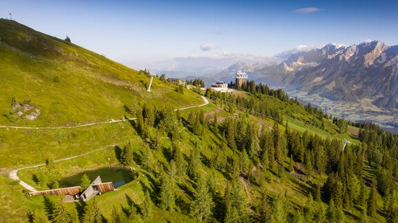 Drohnenaufnahme von der Planai mit grüner Berglandschaft, Wanderwegen, Hütten, Wald und Blick auf das Dachsteinmassiv. | © Josh Absenger