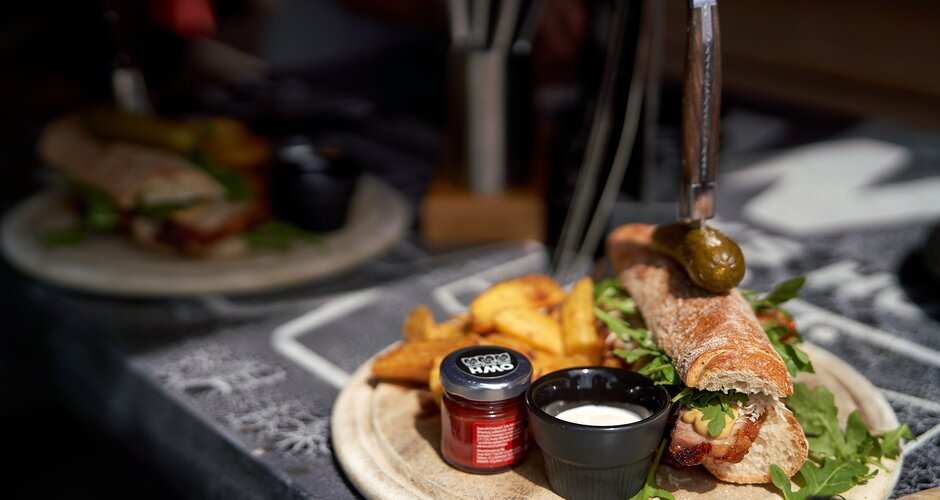 Ein knuspriges Baguette mit Speck, Rucola, Soßen, Essiggurke und Wedges wird auf einer Holzplatte im Sonnenlicht serviert. | © Dietmar Körbler