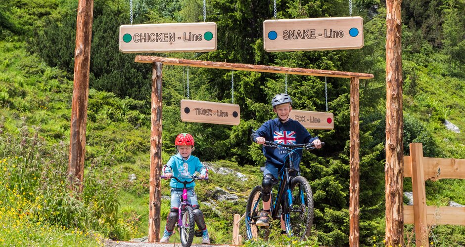 Zwei Kinder fahren mit Mountainbikes in die beschilderten Junior Trails der Reiteralm, umgeben von Blumenwiese und Wald. | © Reiteralm Bergbahnen