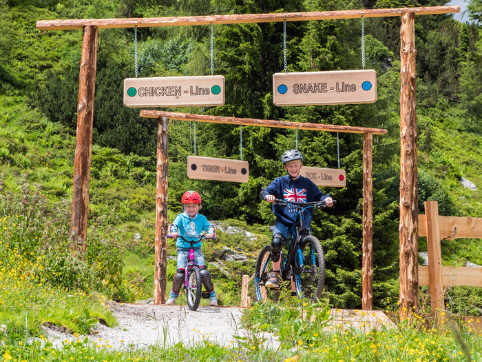 Kinder starten in die Junior Trails auf der Reiteralm bei Sonne | © Reiteralm Bergbahnen Zwei Kinder fahren mit Mountainbikes in die beschilderten Junior Trails der Reiteralm, umgeben von Blumenwiese und Wald. | © Reiteralm Bergbahnen