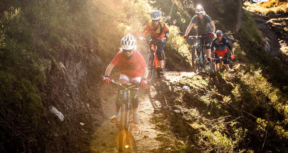 Vier Mountainbiker, darunter zwei Kinder, fahren bei Sonnenlicht auf einem schmalen Trail durch den Wald auf der Reiteralm. | © Reiteralm Bergbahnen, lorenzmasser.com