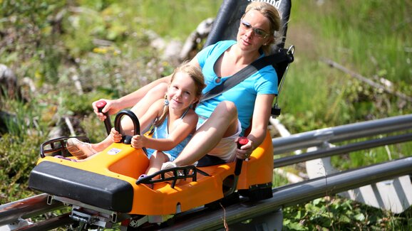 Mutter und Tochter sausen gemeinsam im Sommer mit dem Alpincoaster Rittisberg durch die Kurven, lachend in einem Schlitten. | © photo-austria