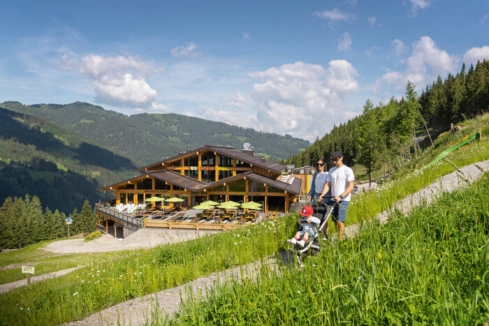 Familienwanderung mit Kinderwagen am Shuttleberg | © Shuttleberg Familie mit Kinderwagen spaziert vor dem Bergrestaurant Lumberjack in grüner Berglandschaft. | © Shuttleberg