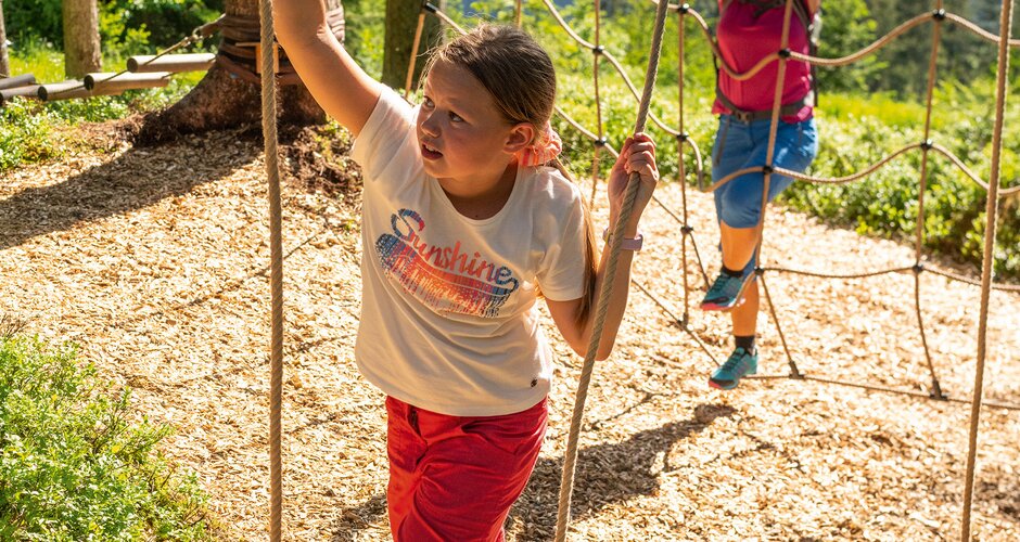 Mädchen balanciert auf Holzsteg im Seilgarten, Frau klettert im Hintergrund. | © Shuttleberg