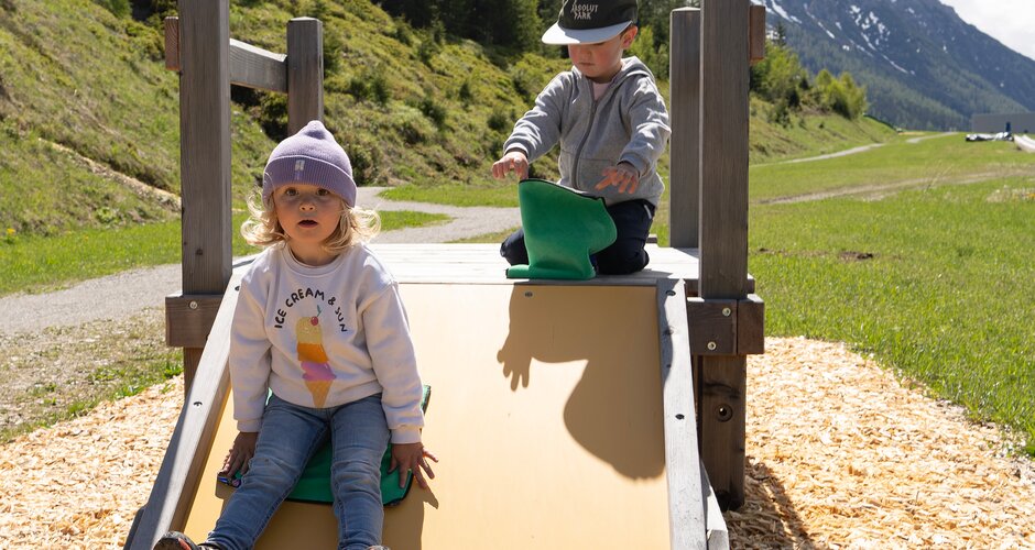Zwei kleine Kinder spielen auf einer flachen Holzrutsche vor grüner Bergkulisse. | © Shuttleberg