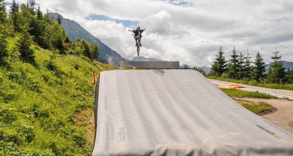 Mountainbiker springt mit ausgebreiteten Armen über Bagjump im Grünen vor Bergkulisse. | © Shuttleberg