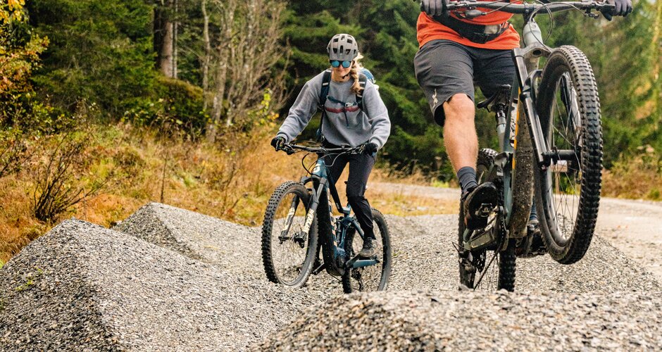 Zwei Mountainbiker fahren über Kieshügel im Wald, einer springt mit dem Bike. | © Shuttleberg