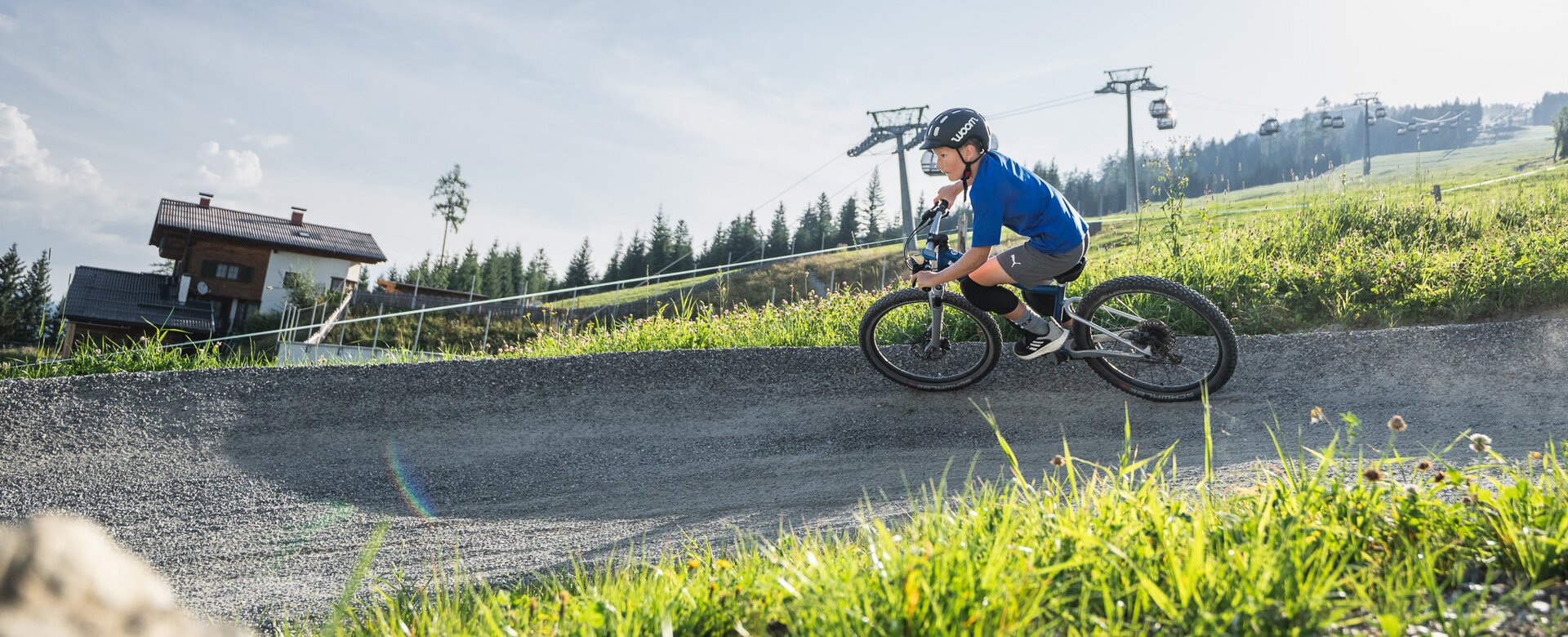 A boy in helmet rides a Woom bike on a curved trail with an alpine house, grassy slope, and gondola lift in the background. | © Snow Space Salzburg - Chris Eder