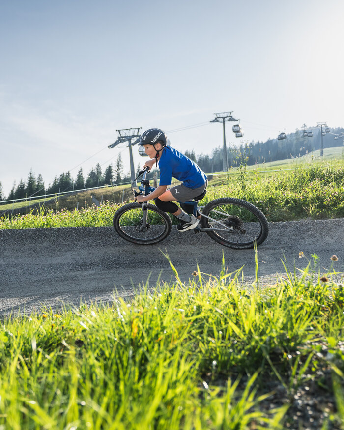 Kind fährt in der woom bike area | © Snow Space Salzburg - Chris Eder Ein Junge mit Helm fährt ein Woom-Bike auf einem Schottertrail, im Hintergrund sind grüne Wiesen, Wald und Gondeln sichtbar. | © Snow Space Salzburg - Chris Eder