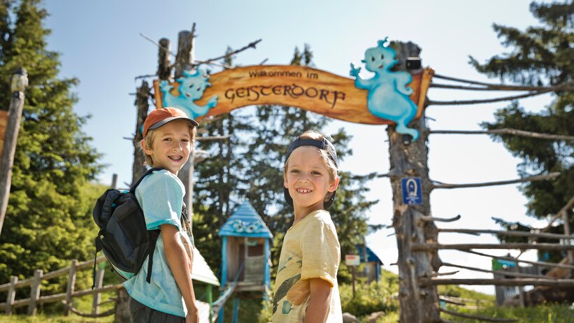 Two smiling boys with backpacks at “Welcome to Ghost Village” sign, colorful wooden huts in the background | © sanktjohann.com, Mirja Geh