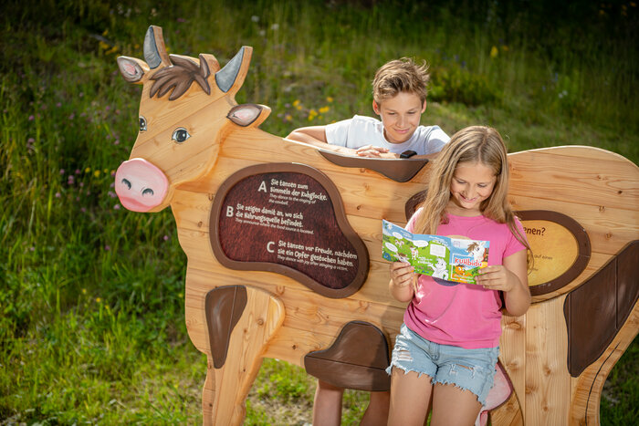 Lernspaß an der KUHbidu-Station mit interaktivem Quiz | © Snow Space Salzburg, Christoph Huber Mädchen liest KUHbidu-Heft, Junge lehnt an hölzerner Kuh mit Quizfrage, grüne Wiese und Blumen im Hintergrund | © Snow Space Salzburg, Christoph Huber