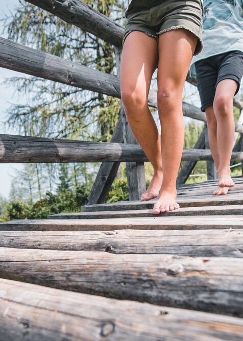 Zwei Personen gehen barfuß über eine Holzbrücke, Nahaufnahme der Beine, im Hintergrund Wiese, Wald und Wanderer | © Snow Space Salzburg
