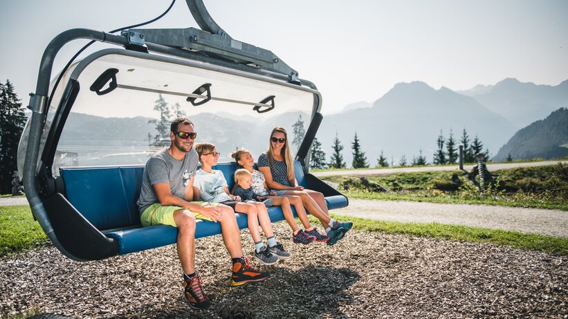 Familie mit drei Kindern sitzt auf Sesselliftbank am Boden, sonniges Wetter, Berge und Nadelbäume im Hintergrund sichtbar | © Snow Space Salzburg