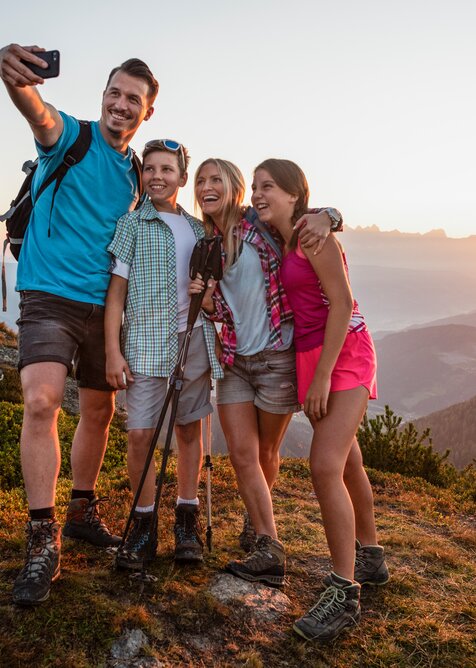 Vierköpfige Familie macht Selfie beim Wandern auf dem Berg, Sonnenuntergang hinter Bergsilhouetten im Hintergrund sichtbar