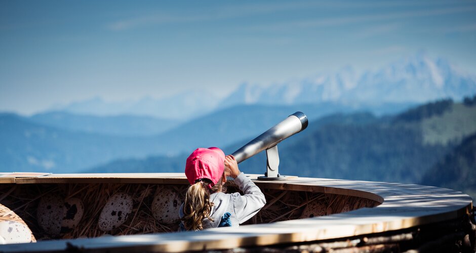 Girl with red cap looks through telescope at alpine panorama from wooden viewing platform in Zauchensee mountains. | © Matthias Fritzenwallner