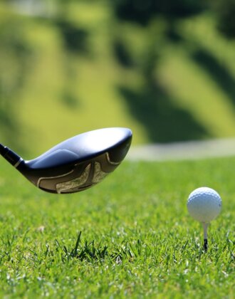 Ein Golfball steht auf einem Tee, kurz vor dem Abschlag mit einem Driver, im Hintergrund ein sonniges Green.