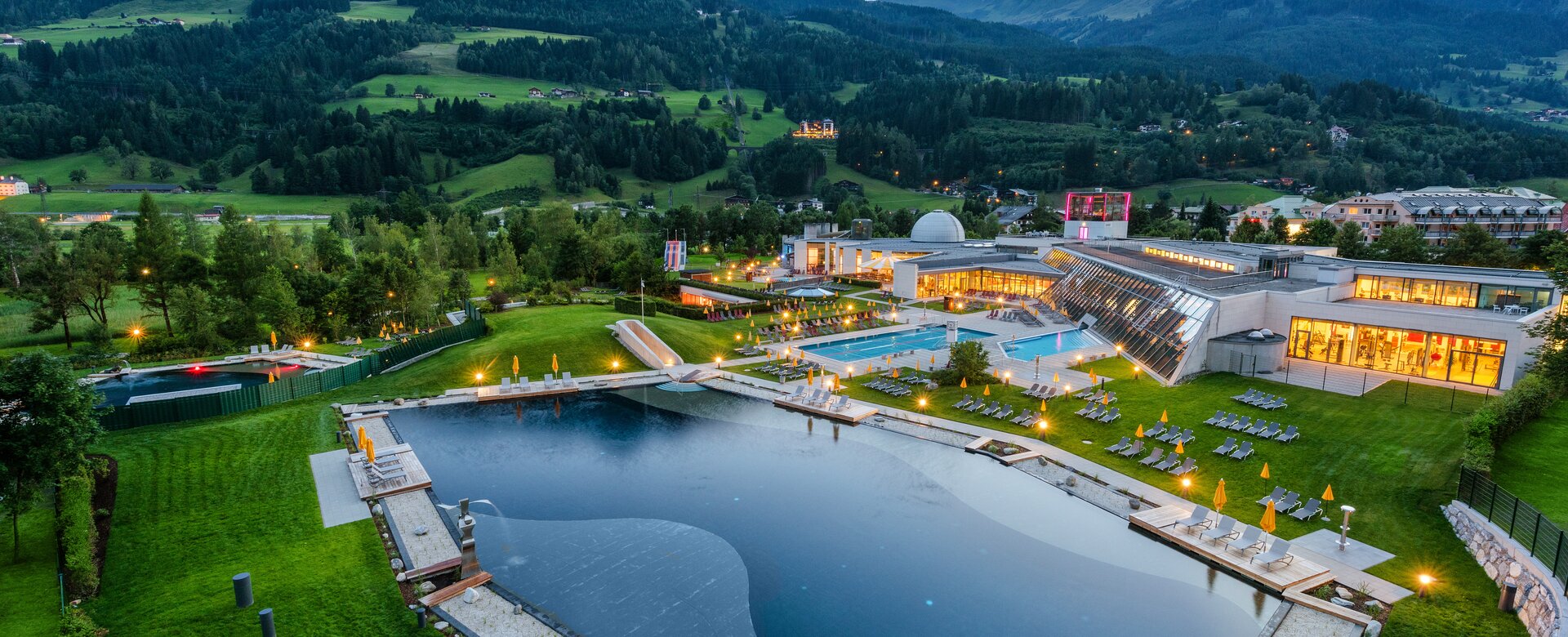 Evening view of Alpentherme Gastein with illuminated bathing lake and green mountain scenery in the background