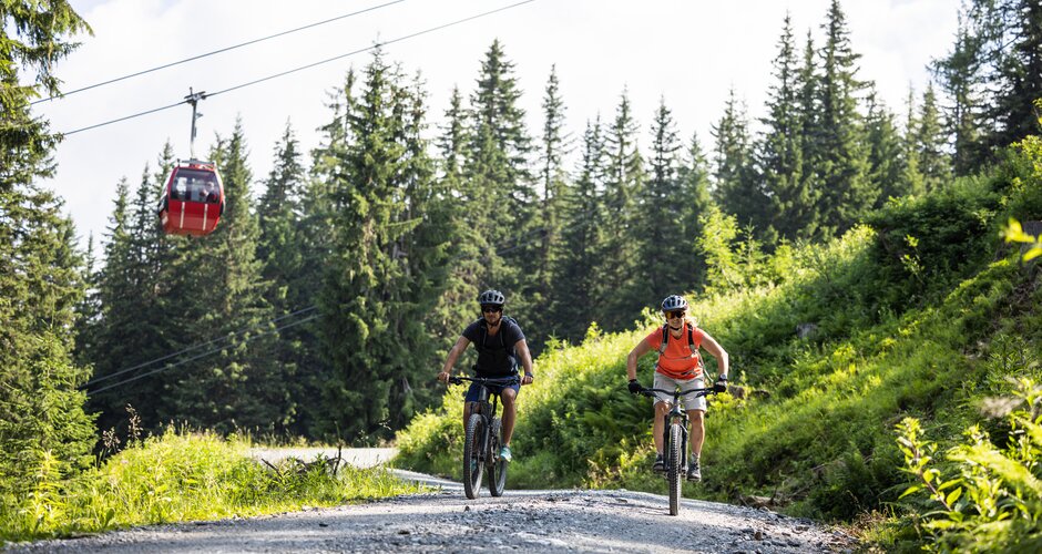 Zwei Mountainbiker auf Schotterweg unter roter Gondelbahn am Fulseck in grüner Berglandschaft | © SalzburgerLand, Heiko Mandl