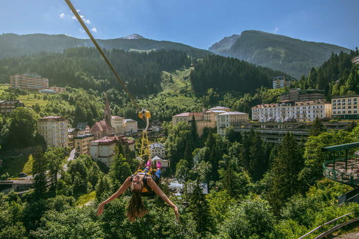 Experience Flying Waters zipline over Bad Gastein | © Max Steinbauer Photography Woman glides on the Flying Waters zipline over rooftops of Bad Gastein, surrounded by forested hills and alpine peaks | © Max Steinbauer Photography