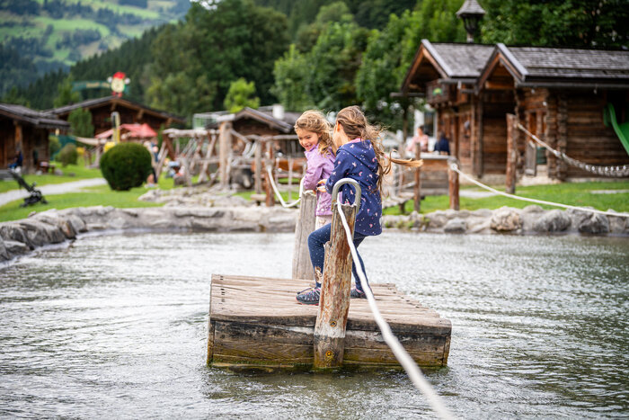 Zwei Kinder spielen auf Holzfloß im Wasser eines Spielplatzes im Großarltal mit Hütten, Wiesen und Bäumen | © Gipfelfieber