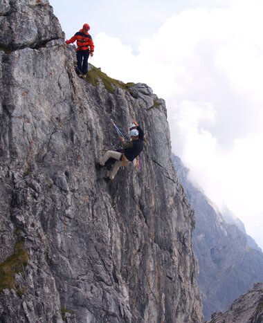 Zwei Kletterer an steiler Felswand gesichert, einer steigt, der andere sichert, Wolken und Bergpanorama im Hintergrund