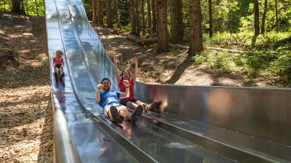 Familie rutscht lachend auf breiter Metallrutsche im Waldrutschenpark durch den schattigen Wald bei Maria Alm | © Roland Haschka