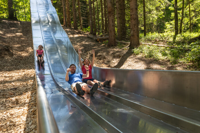 Sliding fun in forest slide park at Natrun, Maria Alm | © Roland Haschka Family laughing while sliding down wide metal slide in forest slide park at Maria Alm's Natrun mountain | © Roland Haschka