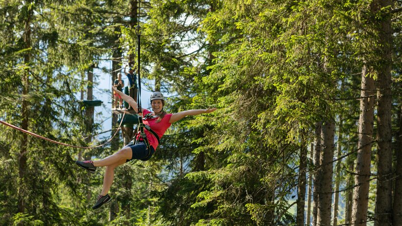 Frau mit Helm und Klettergurt saust lachend am Seil durch den Waldseilgarten Natrun in Maria Alm | © Roland Haschka