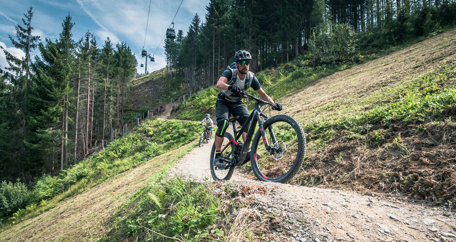 Biker mit Helm und Schutzkleidung fährt auf Flowtrail One in Maria Alm, im Hintergrund Gondelbahn und Wald | © Schartner