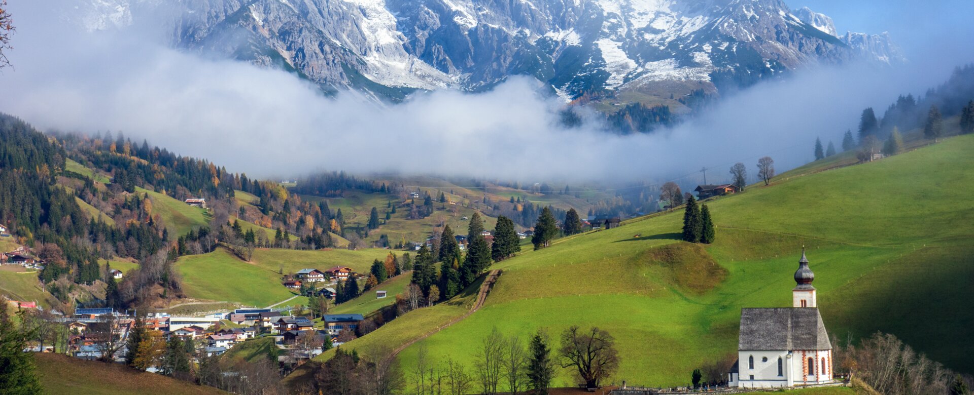 Weiße Kirche bei Dienten auf grünem Hügel, im Hintergrund der verschneite Hochkönig über nebelverhüllten Tälern