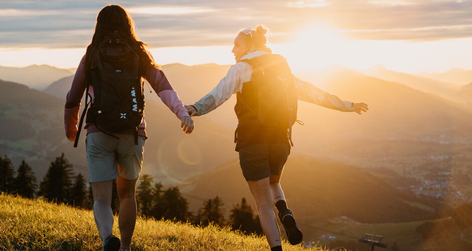 Zwei Frauen mit Rucksäcken gehen über eine Wiese im Abendlicht mit Blick auf Berge und Tal in der Region Hochkönig. | © Hochkönig Tourismus