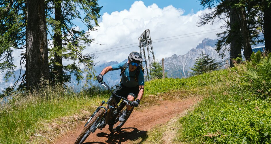 Ein Mountainbiker fährt durch eine Kurve auf einem Dirttrail zwischen Bäumen vor Bergen in der Region Hochkönig. | © Hochkönig Tourismus GmbH