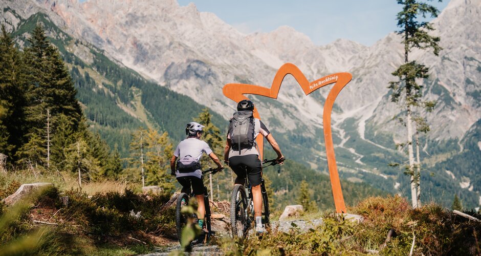 Zwei Mountainbiker fahren auf einem Schotterweg auf einen Torbogen zu, vor hohen Felsbergen in der Region Hochkönig. | © Hochkönig Tourismus GmbH