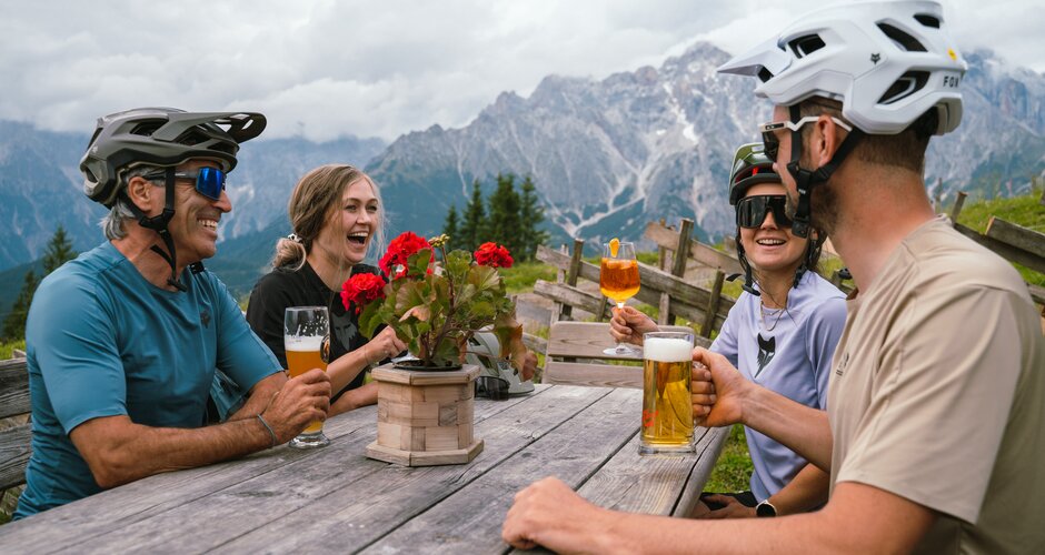 Vier Radfahrer mit Helmen sitzen an einem Holztisch mit Getränken vor Bergen in der Region Hochkönig. | © Hochkönig Tourismus GmbH