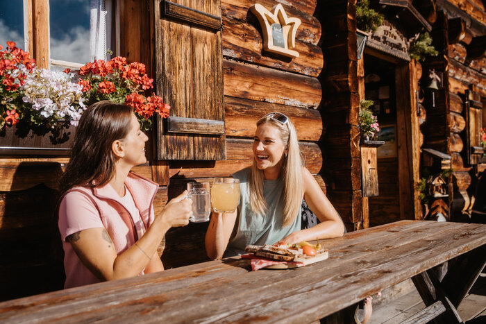Zwei Frauen sitzen an einem Holztisch vor einer Almhütte mit Getränken und Jause im Sonnenlicht am Hochkönig. | © Hochkönig Tourismus GmbH