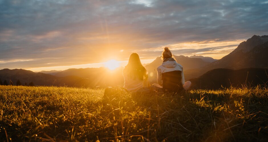 Zwei Frauen sitzen auf einer Wiese und blicken in den Sonnenuntergang über Bergsilhouetten unter wolkigem Himmel. | © Hochkönig Tourismus GmbH