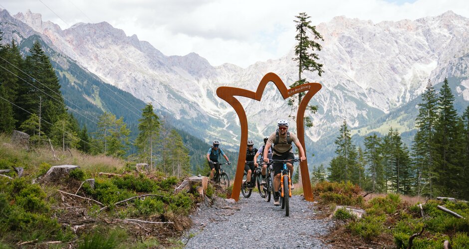 Vier Mountainbiker fahren auf einem Schotterweg durch einen Torbogen vor Felsbergen und Tannen in der Region Hochkönig. | © Hochkönig Tourismus GmbH