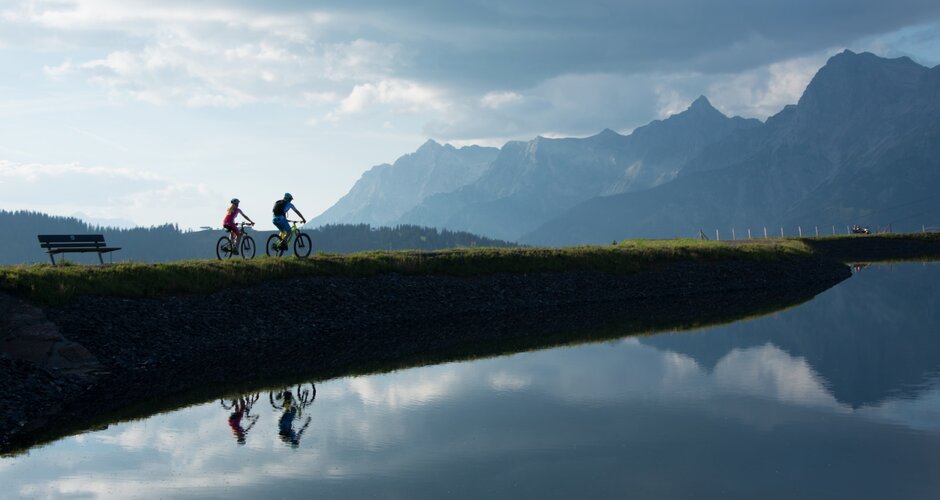 Zwei Mountainbiker fahren am ruhigen Speichersee entlang, Bergsilhouette Hochkönig spiegelt sich im Wasser | © Hochkönig Tourismus, Schartner