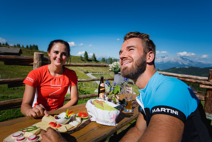 Genuss auf der Alm: Jause mit Ausblick in den Alpen | © Salzburger Sportwelt - Christian Schartner Mann und Frau genießen bei Sonnenschein eine Almjause mit Käseplatte, Brot und Getränken vor Alpenkulisse in der Salzburger Sportwelt | © Salzburger Sportwelt - Christian Schartner